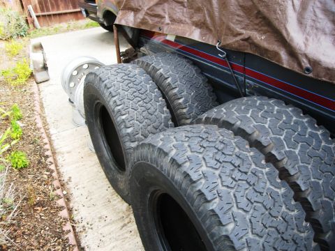 1986 Ford Bronco - A collection of off-road tires and chrome wheels are visible, partially obscuring a tarp-covered vehicle.