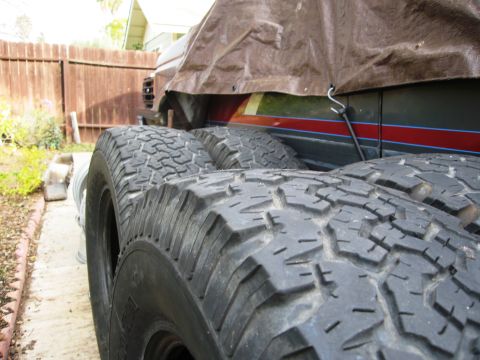 1986 Ford Bronco - Tires are in the foreground, with the side of a covered vehicle visible behind them.