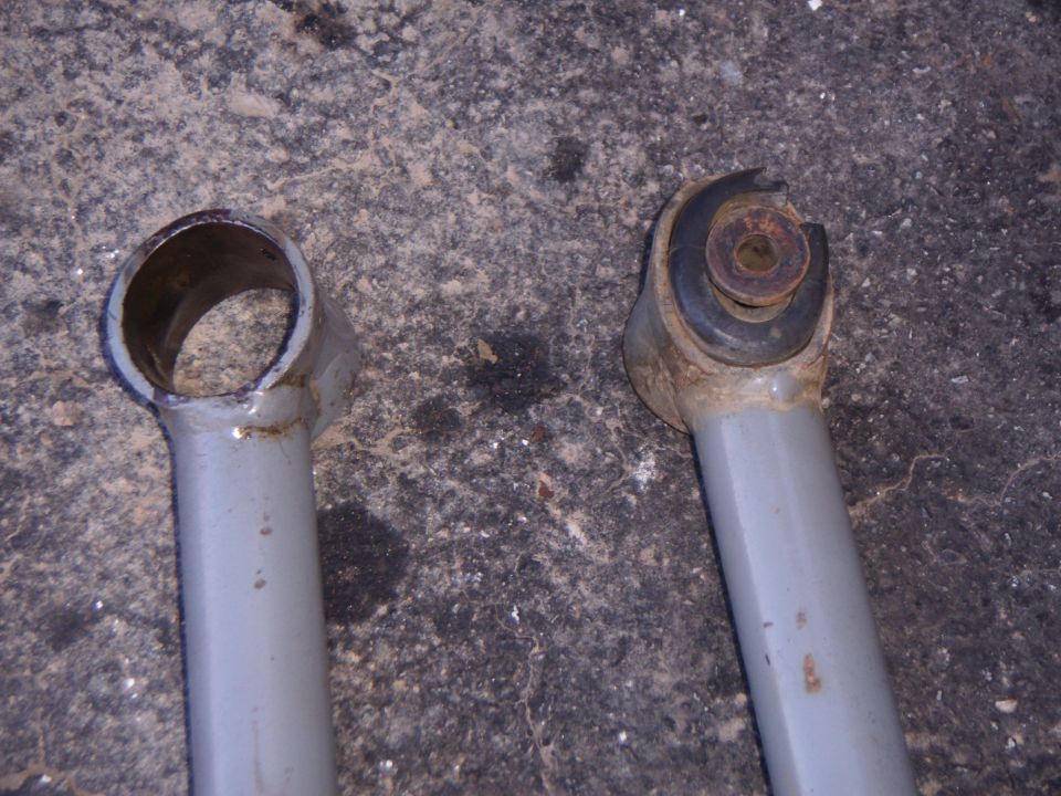 Two gray tubular control arm ends on concrete, one empty eye and one with a rusty bushing