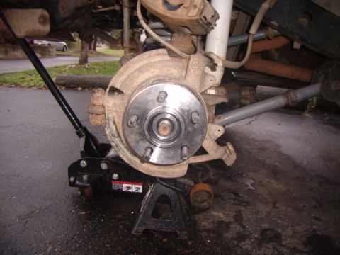 2003 Jeep Wrangler - Close-up view of a vehicle's front wheel hub assembly, supported by a jack and jack stand, with the brake rotor and lug nuts visible.