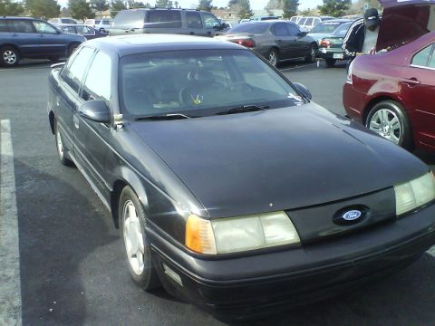 1993 Ford Taurus - A dark sedan is parked in a lot with other vehicles around it.