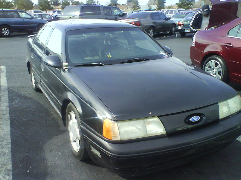 Dark sedan parked in a lot, front end visible. Another car's trunk is open nearby
