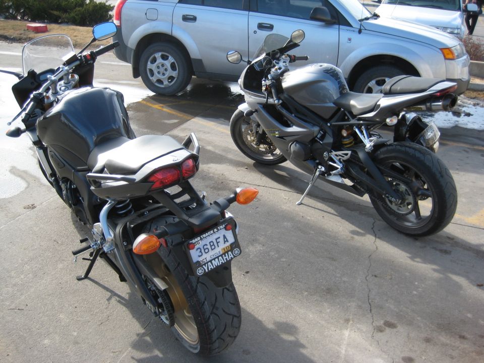 Two motorcycles parked on asphalt with a silver SUV behind them. The motorcycle on the left is black, and the one on the right is dark gray