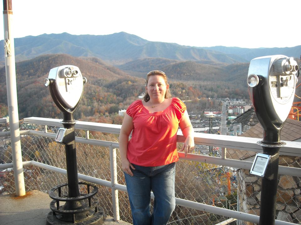 Woman posing for a photo on an observation deck with mountains and a town in the background. Two coin-operated binoculars are visible