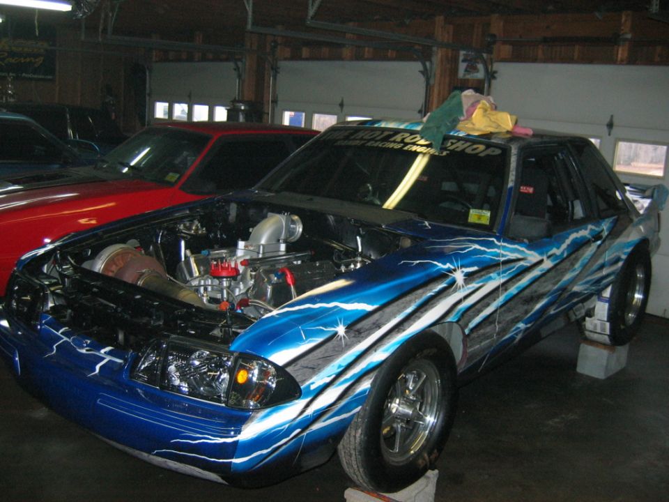 Blue race car with lightning graphics, hood open showing engine and turbo. Red car behind it