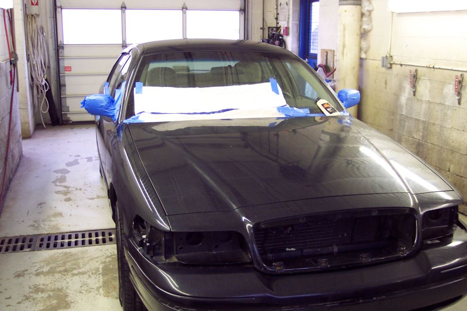 Dark sedan with blue tape on side mirrors and windshield, missing headlights, in a garage