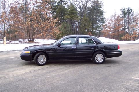 2001 Ford Crown Victoria - Side profile of a black 2001 Ford Crown Victoria sedan parked in a lot with trees and snow in the background.