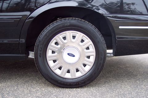 2001 Ford Crown Victoria - The rear wheel and fender of a black 2001 Ford Crown Victoria are visible, a chrome hubcap with the Ford logo.
