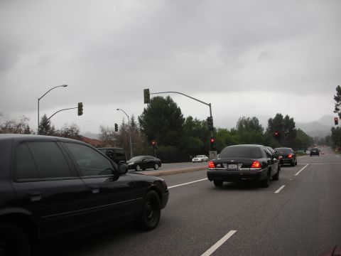 1992 Ford Crown Victoria - Two black Crown Victoria police cars are driving on a road under a cloudy sky.