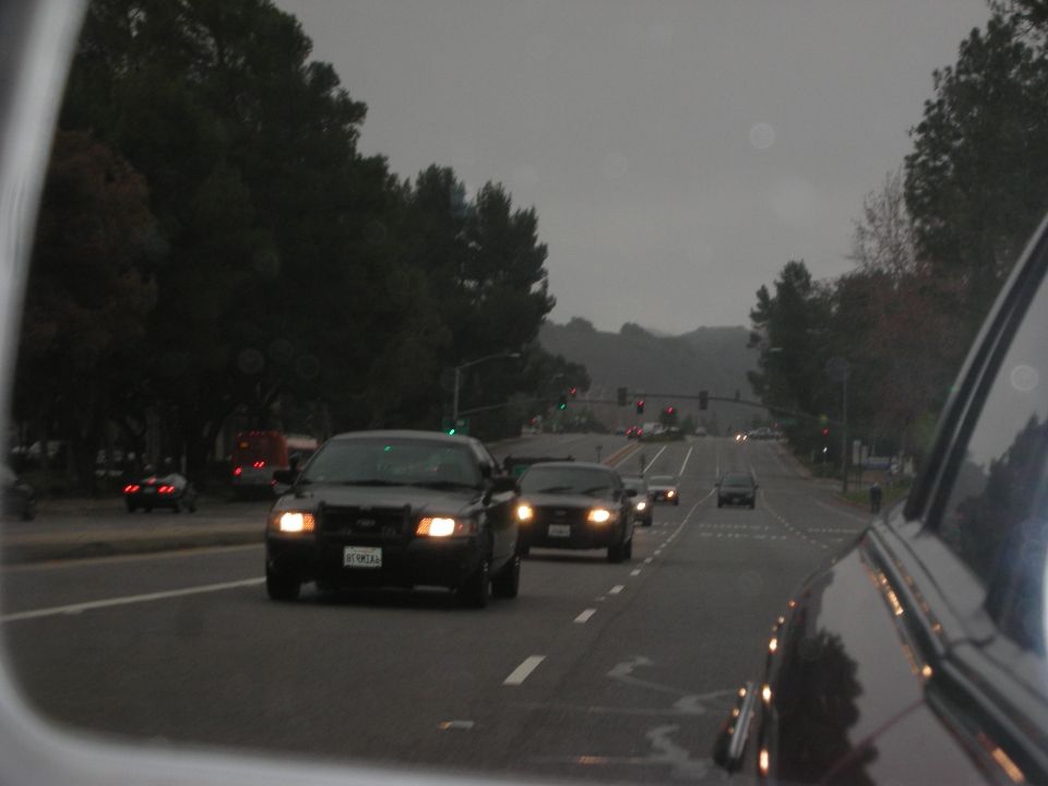 Black police car with headlights on, license plate visible, driving on a wet road