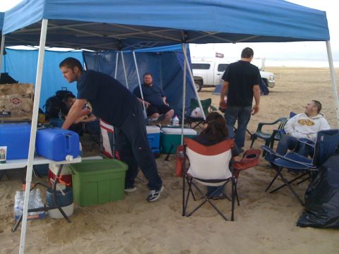 1989 Ford Bronco - A white pickup truck is visible behind a group of people gathered under a blue canopy on a beach.