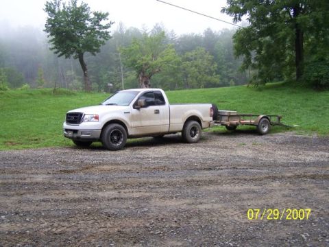 2004 Ford F-150 - A white 2004 Ford F-150 pickup truck is parked on a gravel surface, attached to a utility trailer, with a wooded area in the background.