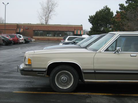 1988 Mercury Grand Marquis - A 1988 Mercury Grand Marquis sedan is parked in a lot with other vehicles, with a building in the background.