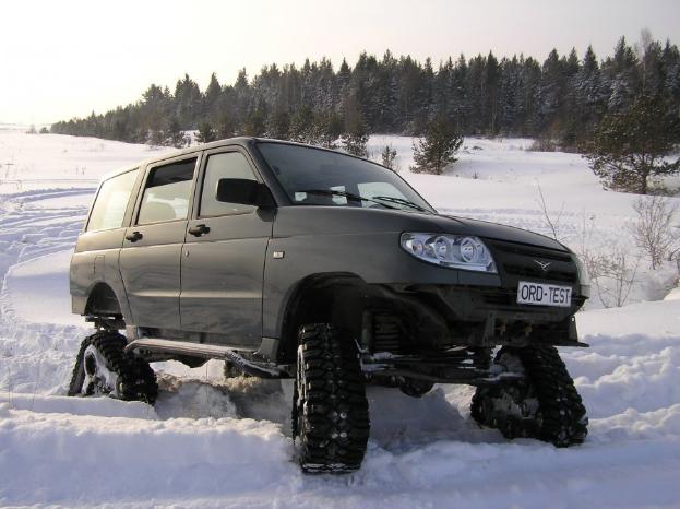 Black SUV fitted with rubber track systems parked in deep snow before a pine forest.