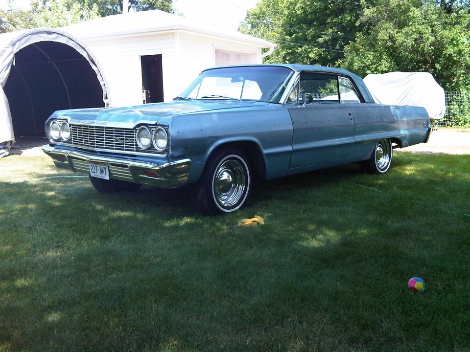 1964 Chevrolet Impala coupe parked on grass in front of a garage and a covered structure