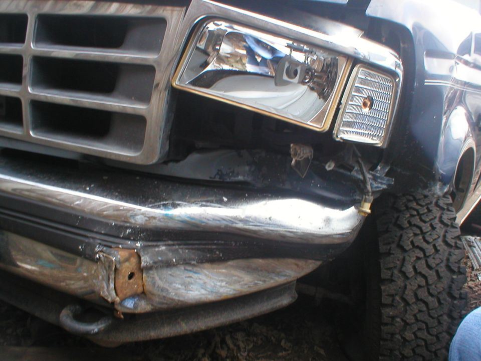 Front corner of dark truck with crushed chrome bumper and dented fender after a collision