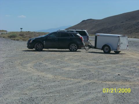 2009 Kenworth T800 - A dark SUV is seen towing a white trailer on a gravel road under a clear blue sky.