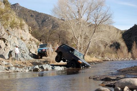 1996 Ford Bronco - Two off-road vehicles navigate a shallow riverbed with rocky terrain and sparse trees.