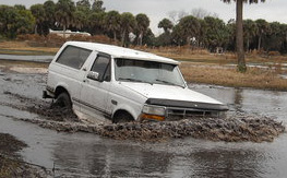1992 Ford Bronco - A white 1992 Ford Bronco struggles through deep mud and water, with palm trees and overcast skies in the background.
