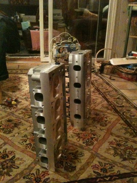 1972 Ford Ranchero - Two freshly machined aluminum cylinder heads standing on end on a floral area rug in a living room, with intake/exhaust ports and bolt hole…