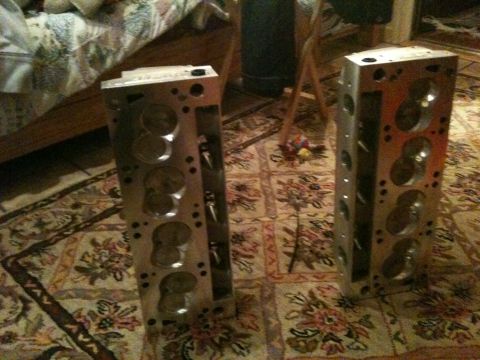 1972 Ford Ranchero - Two bare aluminum V8 cylinder heads with no valvetrain installed stand upright on end on a floral area rug indoors, ports and bolt holes ex…