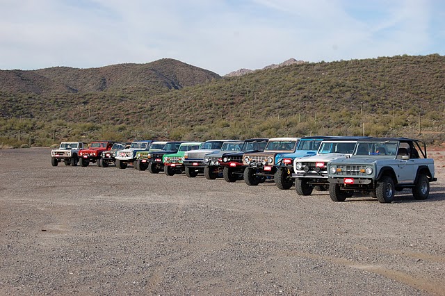 Line of vintage Ford Broncos in various colors parked on gravel with mountains in the background