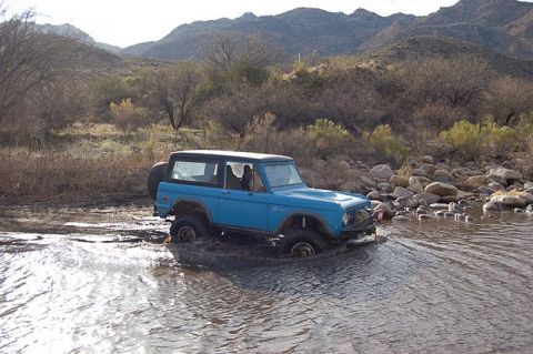 1996 Ford Bronco - First-generation Ford Bronco navigates shallow water and rocky terrain in a desert landscape.
