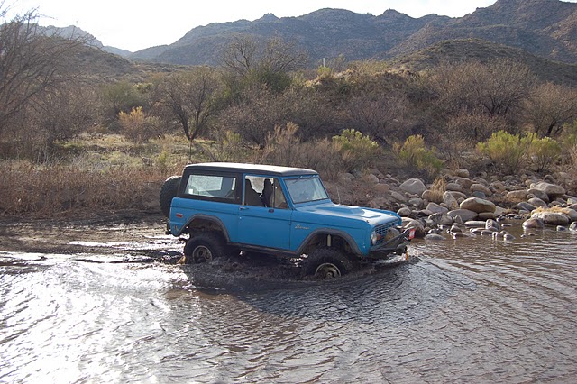 Blue first-generation Ford Bronco driving through a rocky stream bed