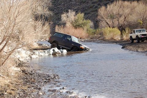 1996 Ford Bronco - A dark Ford Bronco is stuck in a shallow river, while another Bronco waits on the bank.