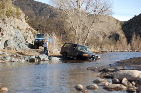 1996 Ford Bronco - Two SUVs navigate a shallow river crossing in a rocky, arid landscape.