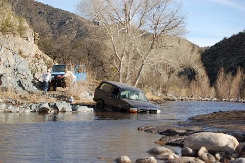1996 Ford Bronco - A dark SUV is stuck in a river while two people stand by another vehicle on the rocky shore.