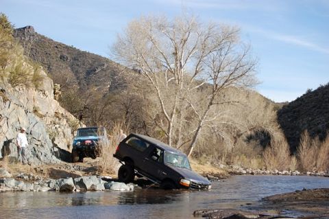 1996 Ford Bronco - A dark blue full-size Ford Bronco tips steeply nose-down off a rocky ledge at the edge of a shallow desert river, with a driver inside and…