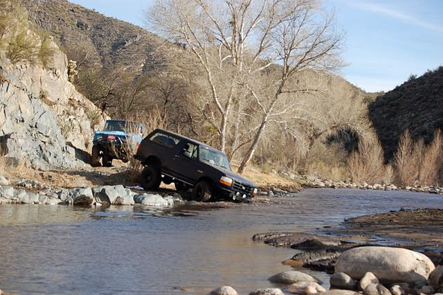 Dark blue Ford Bronco dropping off a rock ledge into a shallow desert river