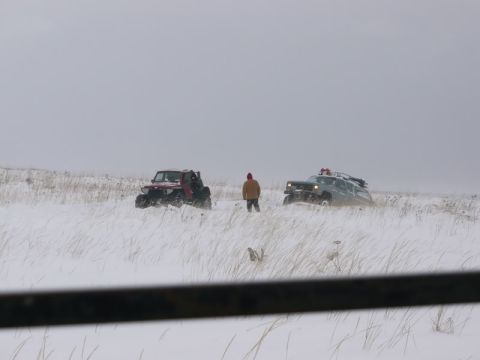 1987 Suzuki Samurai - A person stands between two off-road vehicles in a snow-covered field, with dry grass poking through the snow.