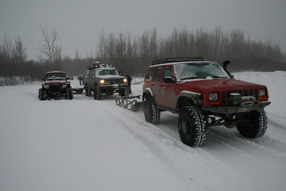 Three modified off-road vehicles driving on a snowy path. The closest is a red Jeep Cherokee with a winch. Another vehicle is towing a sled