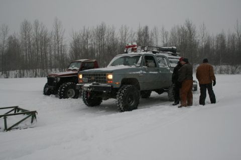 1987 Suzuki Samurai - Two off-road trucks are parked in a snowy landscape with bare trees in the background.