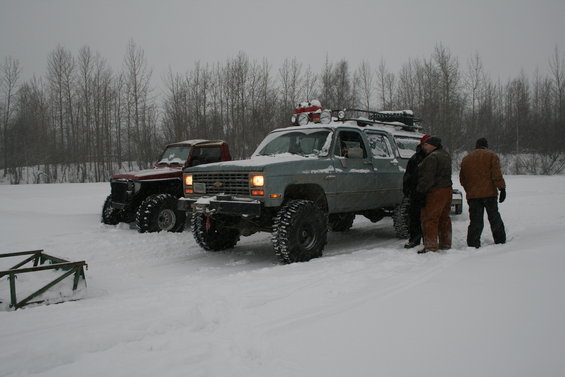 Two modified trucks with large tires sit in the snow. One is a gray SUV, the other a red pickup
