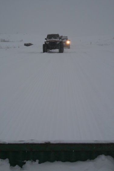 1987 Suzuki Samurai - Two off-road vehicles navigate a snowy landscape, their headlights cutting through the overcast sky.