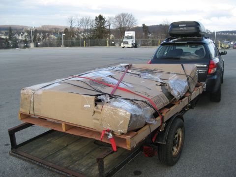 1996 Ford Bronco - A car tows a trailer loaded with a large, strapped-down box, with a semi-truck visible in the distance.