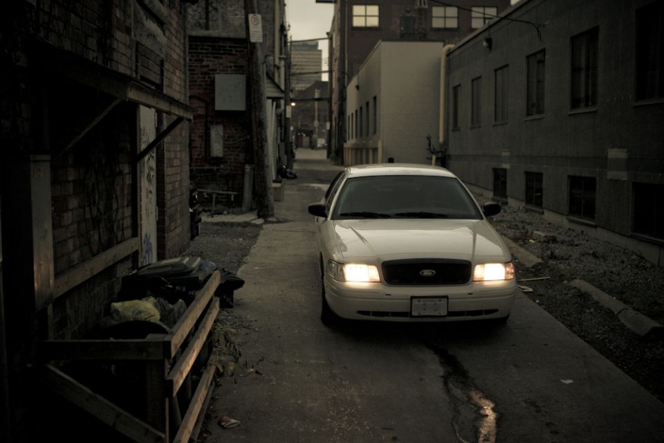 White Ford Crown Victoria sedan with headlights on in a dark urban alley