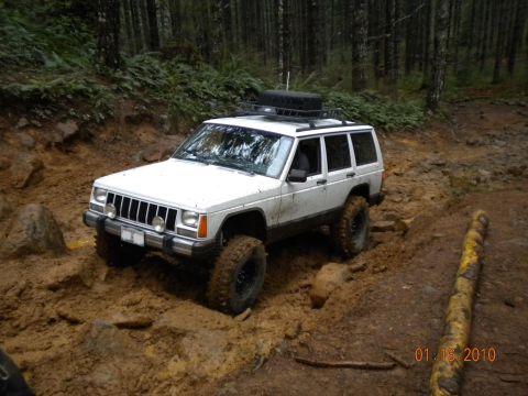 2005 Jeep Wrangler - White Jeep Cherokee XJ driving through a muddy forest trail with large off-road tires.