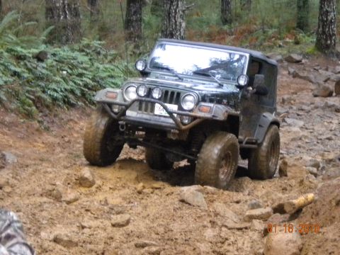 2005 Jeep Wrangler - A heavily lifted dark Jeep Wrangler TJ flexes through a muddy, rocky trail section, front axle articulated with one tire dropped into a rut…