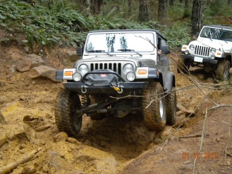 2005 Jeep Wrangler - A khaki-gold Jeep Wrangler Rubicon with black soft top flexes its front axle through a deep dirt rut, showing the covered Warn winch, steel…