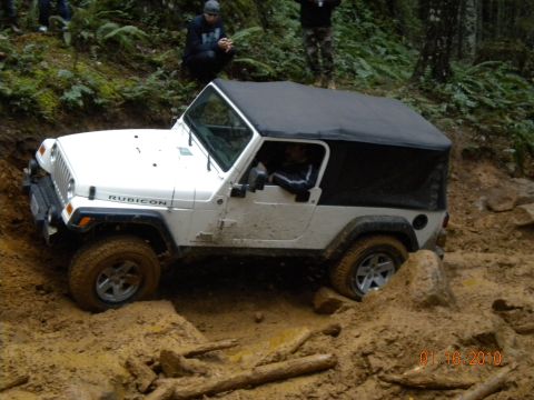 2005 Jeep Wrangler - A white long-wheelbase Jeep Wrangler Unlimited Rubicon with a black soft top works through a deeply rutted mud pit on a wet forest trail, m…