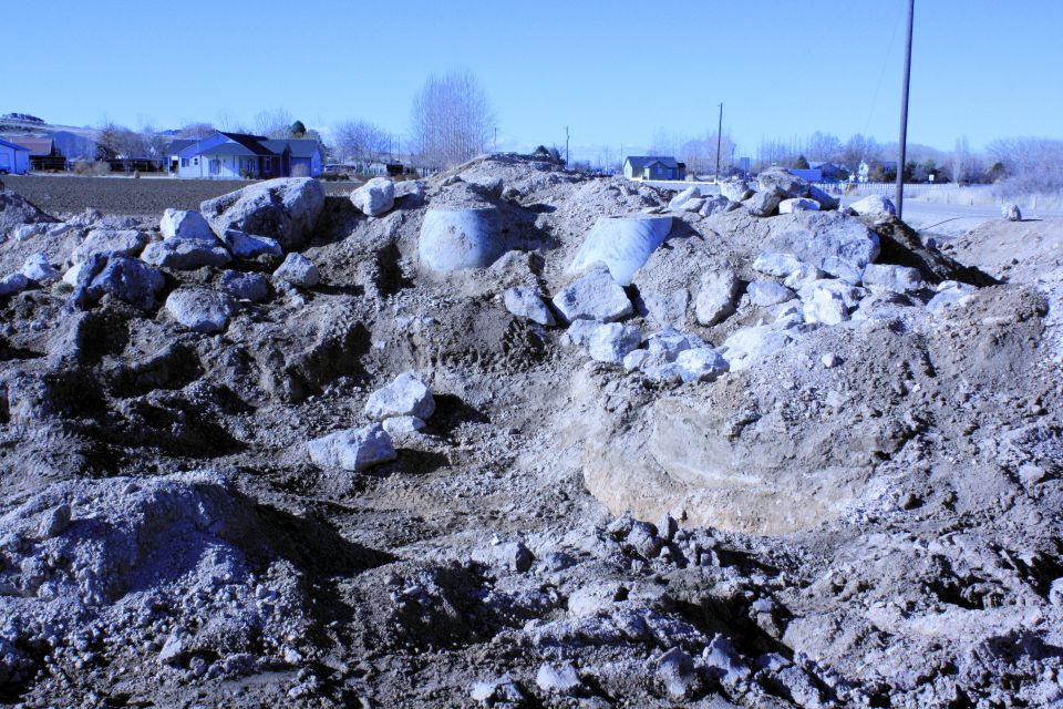 Pile of dirt, rocks, and concrete pipes at a construction site with houses in the background
