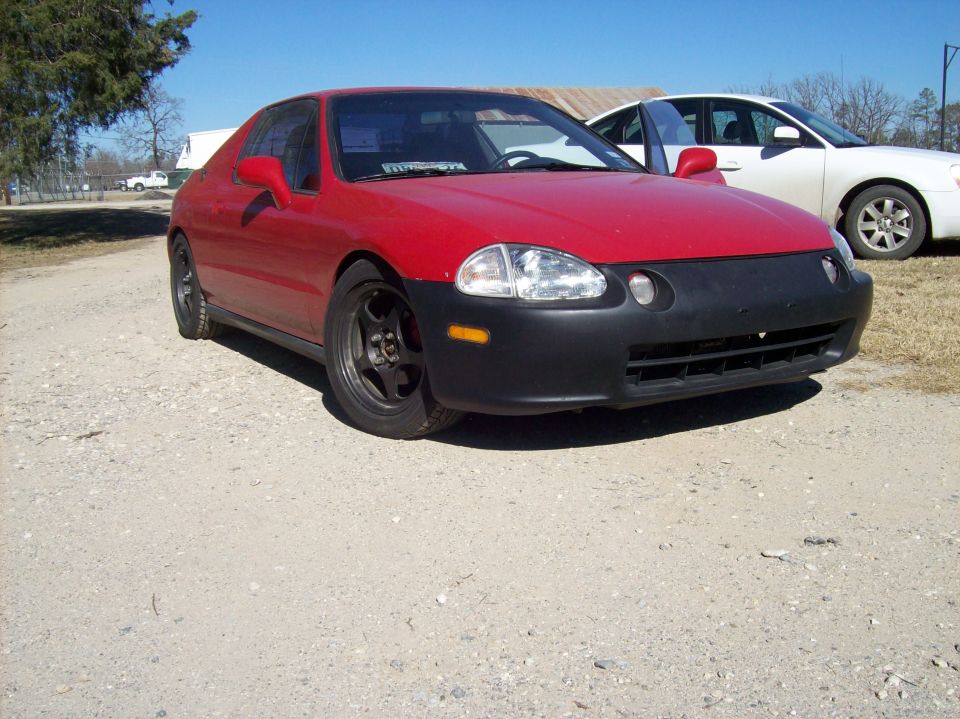 Red Honda del Sol with black front bumper and aftermarket wheels, parked on a gravel lot. White sedan in background