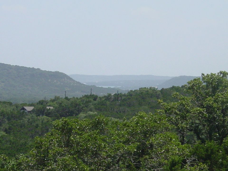 Hazy hill country landscape with dense green trees, distant lake, and a rooftop among foliage