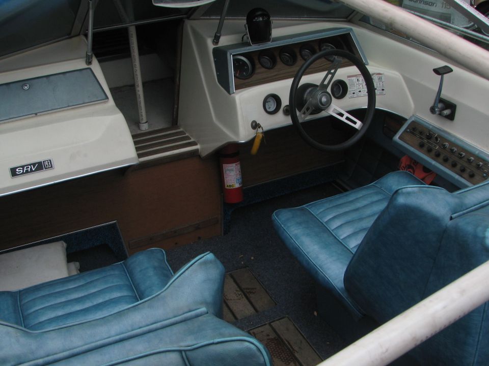 Boat interior with blue seats, dashboard with gauges, and steering wheel. Fire extinguisher visible
