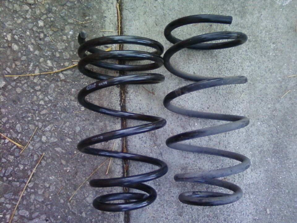 Two black coil springs on concrete, an Eibach spring next to a stock coil spring