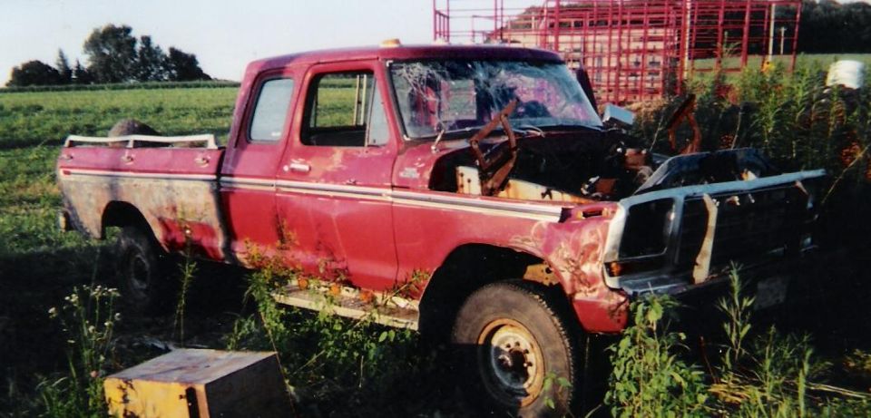 Wrecked red Ford pickup in weeds with missing hood, smashed windshield, and detached grille
